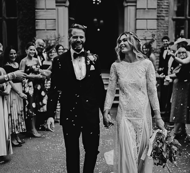 Bride in a high neck long sleeve boho lace wedding dress walks with the groom in black tie as they have their confetti moment 