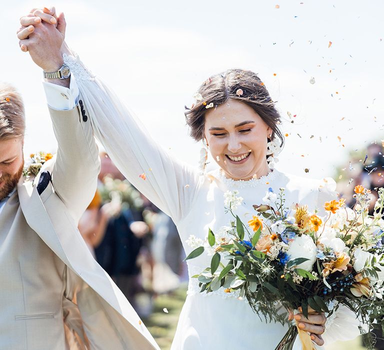 Groom in cream wedding suit with a dark blue and handkerchief lifting his hand with the bride in a vintage dress as they walk through confetti 