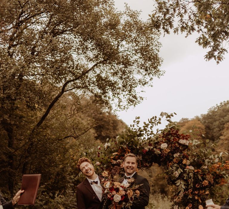 Groom in black tie with groom in a burgundy suit stand at at altar fr their outdoor ceremony with a warm orange bouquet 