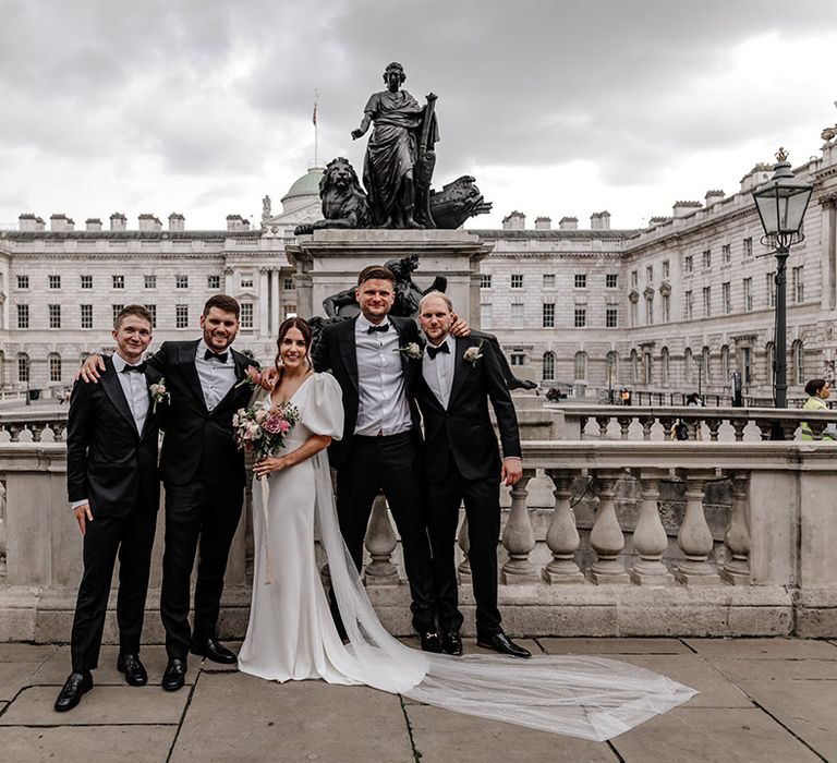 Bride in modern front slit wedding dress standing with the groomsmen in matching black tuxes at Somerset House