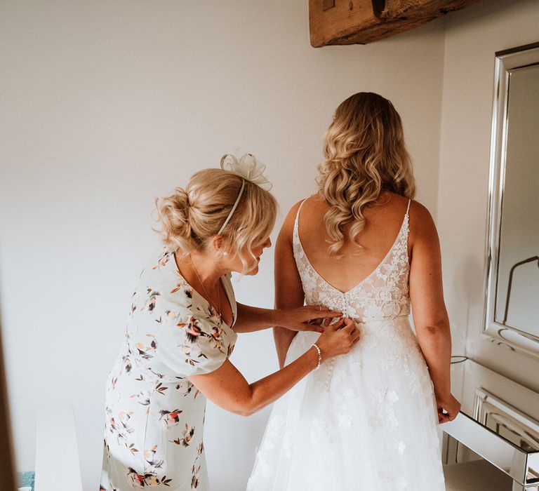 Bride in a low back floral lace wedding dress is buttoned up by the mother of the bride in a floral dress 