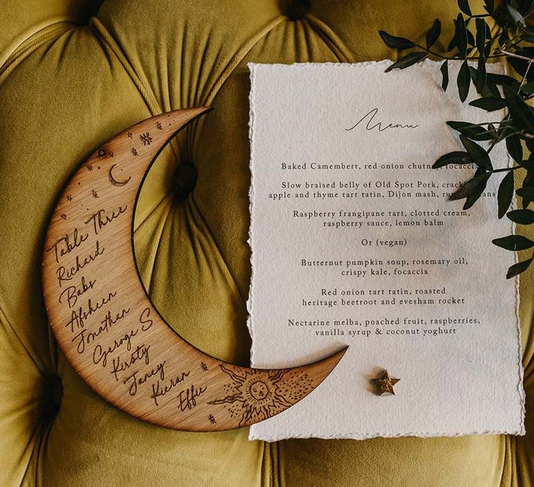 Handmade wooden moon table number with illustrations of the sun and the moon next to minimalistic white wedding menu on olive green velvet sofa