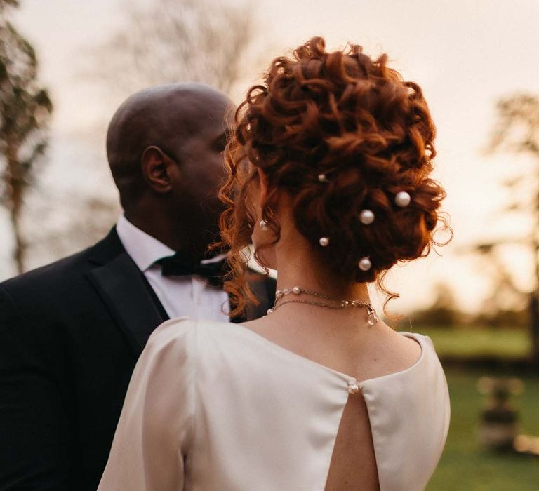 Groom in black tux with bowtie embracing bride in open back detail satin wedding dress with pearl hair accessories and pearl necklace