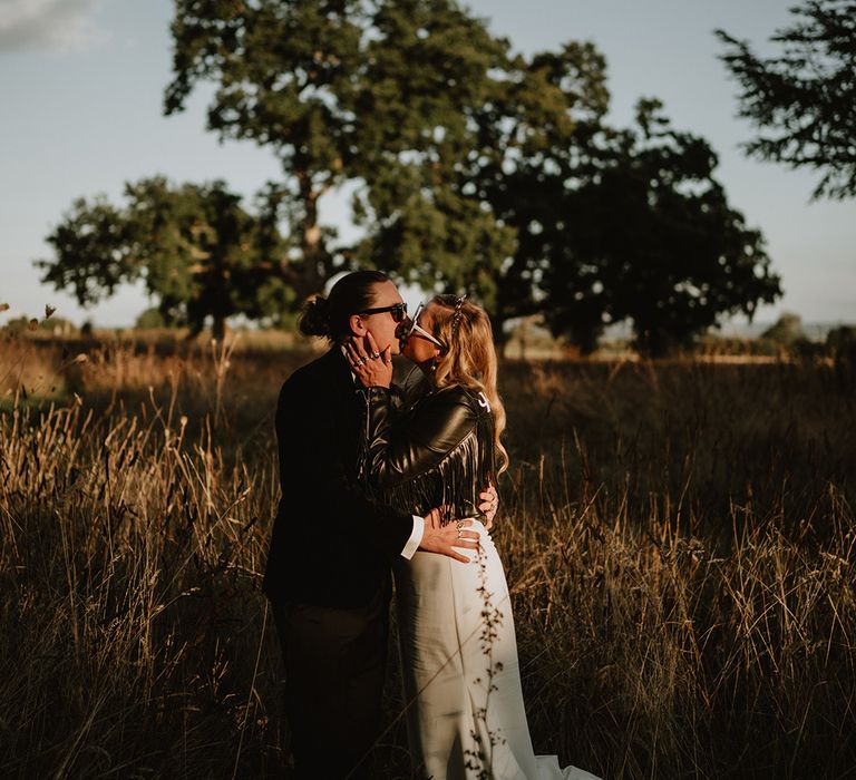 Bride in personalised leather jacket with trimmings kissing groom in dark suit and bow tie, both wearing sunglasses at 70's-inspired wedding with disco balls and backdrop banner