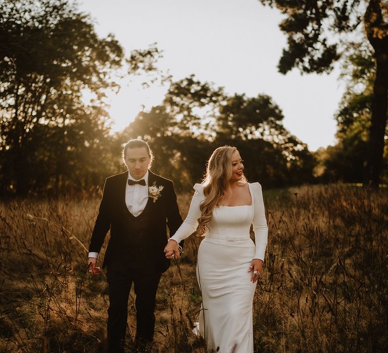 Bride in square neck long sleeve satin wedding dress with two tier veil holding hands with groom in black suit with floral boutonniere walking around St Tewdrics House