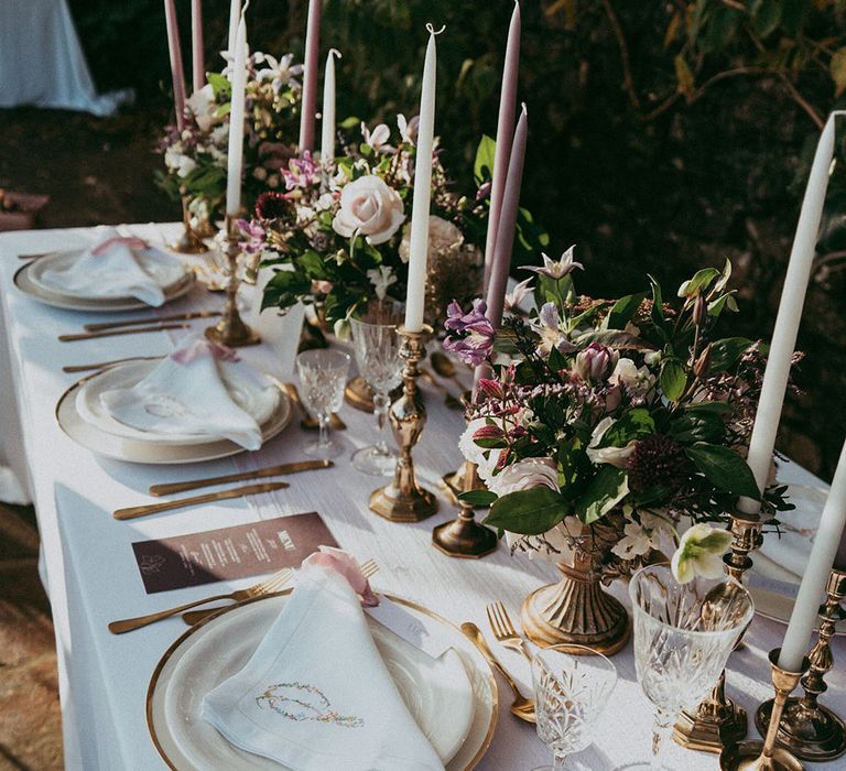 Wedding tablescape with lavender and white tapered candles in gold candle holders, mixed lavender, neutral and white bouquets, embroidered white napkins and gold cutlery 