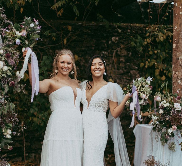 Bride in sleeveless tulle layered wedding dress with v neck detail and bride in short puff sleeve lace wedding dress holding matching bouquets with lavender lace ribbon