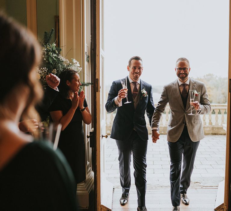 The two grooms enter their wedding reception with glasses of champagne 