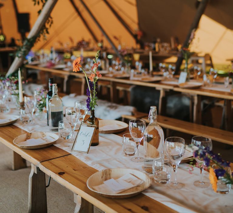 Rustic wooden banquet tables with white table runner and small colourful floral arrangements in tent 