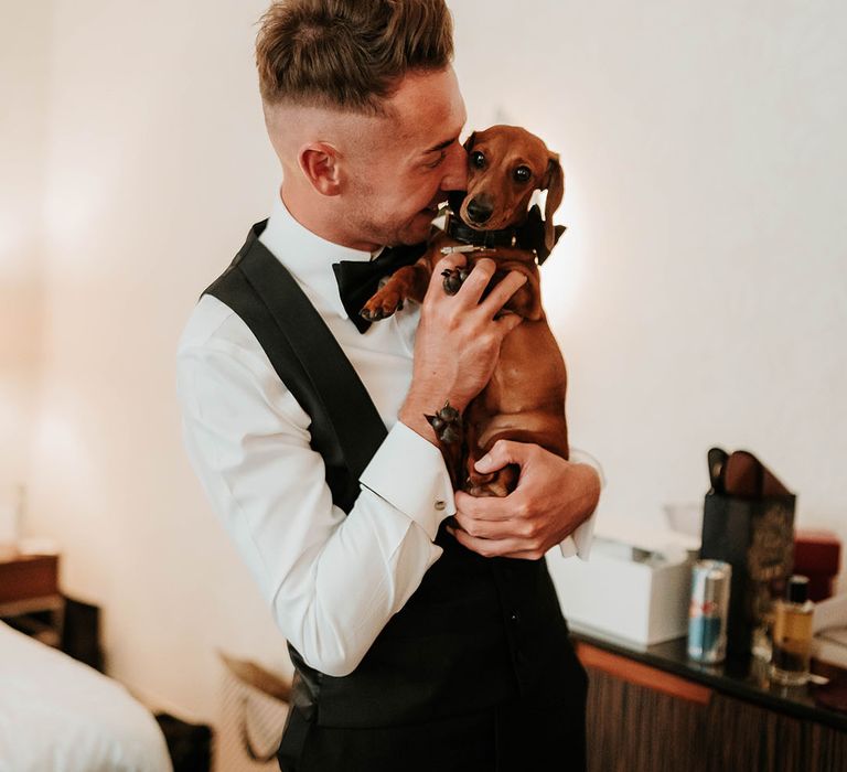 Groom in black waistcoat, black bowtie and crisp white shirt holding sausage dog, getting ready for stylish wedding