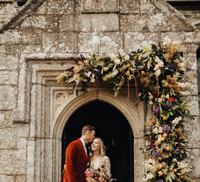 Groom in orange velvet blazer kisses the bride on the forehead as they stand at the entrance to the church 