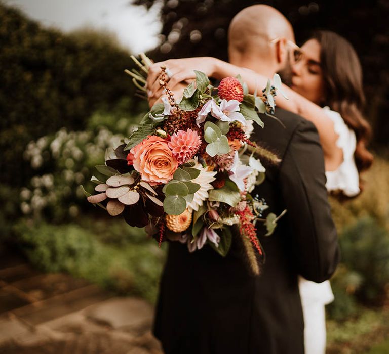 Bride and groom kissing, showing off luxe wedding bouquet with white and pink carnations, garden roses, eucalyptus, baby’s-breath and dried flowers