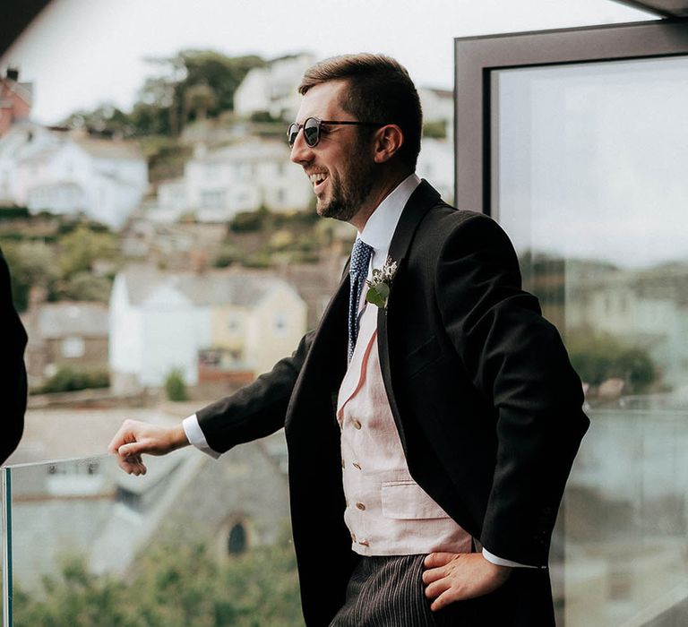 Groom in sunglasses, pale pink waistcoat and blue tie looks out across to the sea for Salcombe wedding 