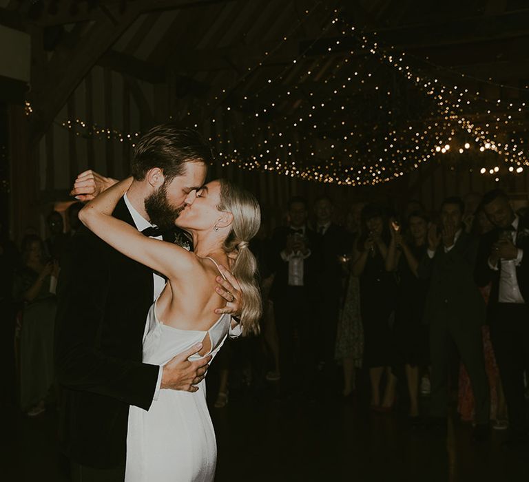 The bride and groom share a passionate kiss as they participate in their first dance together under twinkling fairy lights 