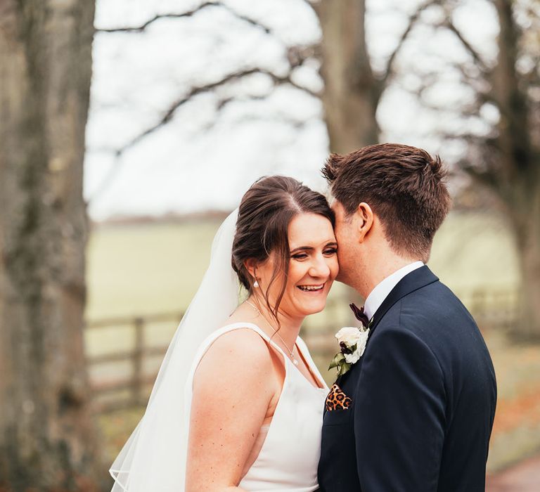 Groom in a navy suit with a leopard print pocket square embracing the bride in a Maggie Sottero wedding dress with pearl jewellery for a Christmas wedding 