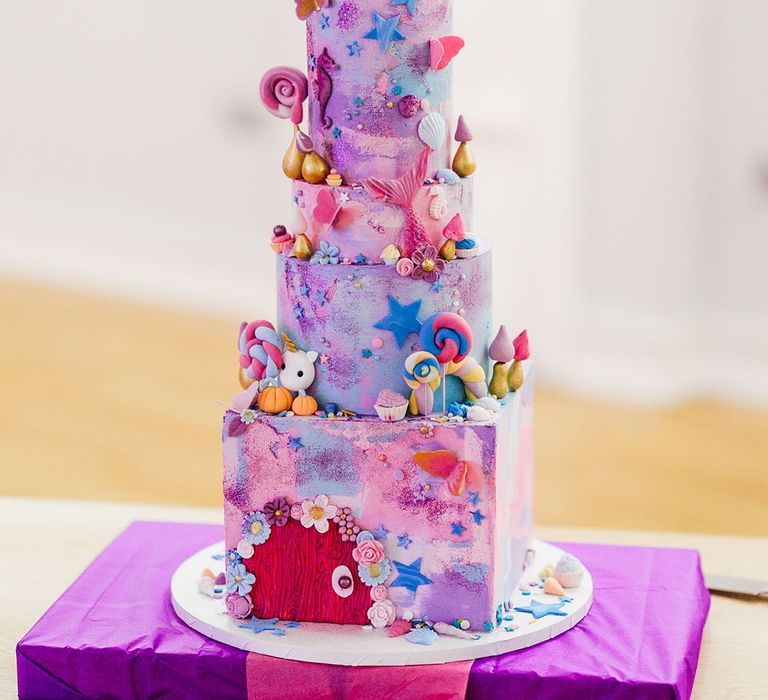 Four tier pink and purple iced wedding cake with cartoon and fairy decorations, lollipop, star, and flower icing and personalised pink and blue wedding cake toppers 