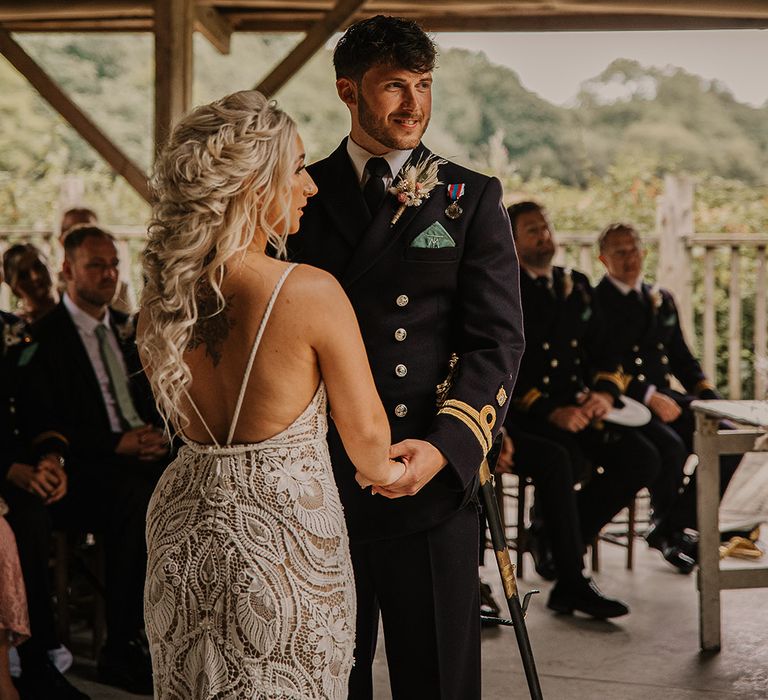 Bride in open back boho lace wedding dress with hair in a fishtail braid standing with the groom in a military uniform at the altar for their ceremony 