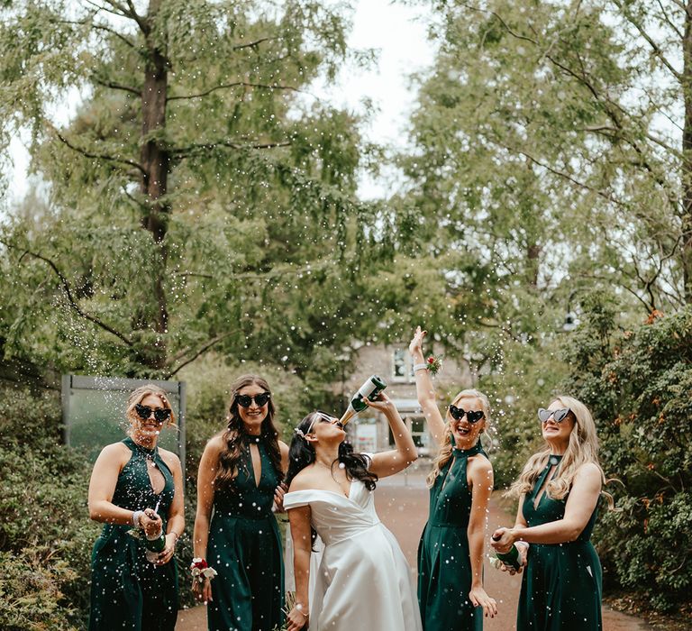 Bride stands between her bridesmaids wearing high-neck green jumpsuits and heart shaped sunglasses whilst spraying champagne 