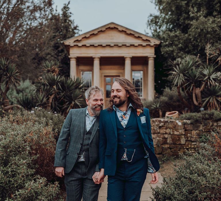 Groom in three piece tartan suit and groom in blue three piece tartan suit walk alongside one another at the Kew Gardens 