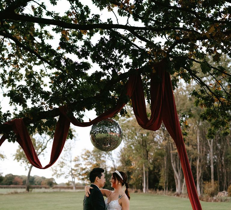 Bride & groom kiss beneath disco ball decor outdoors finished with burnt orange drape across tree 