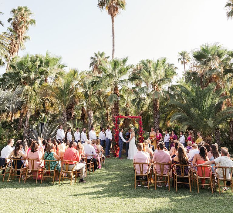 Brides stands to the front of aisle beneath red floral arch during outdoor wedding ceremony in Mexico 