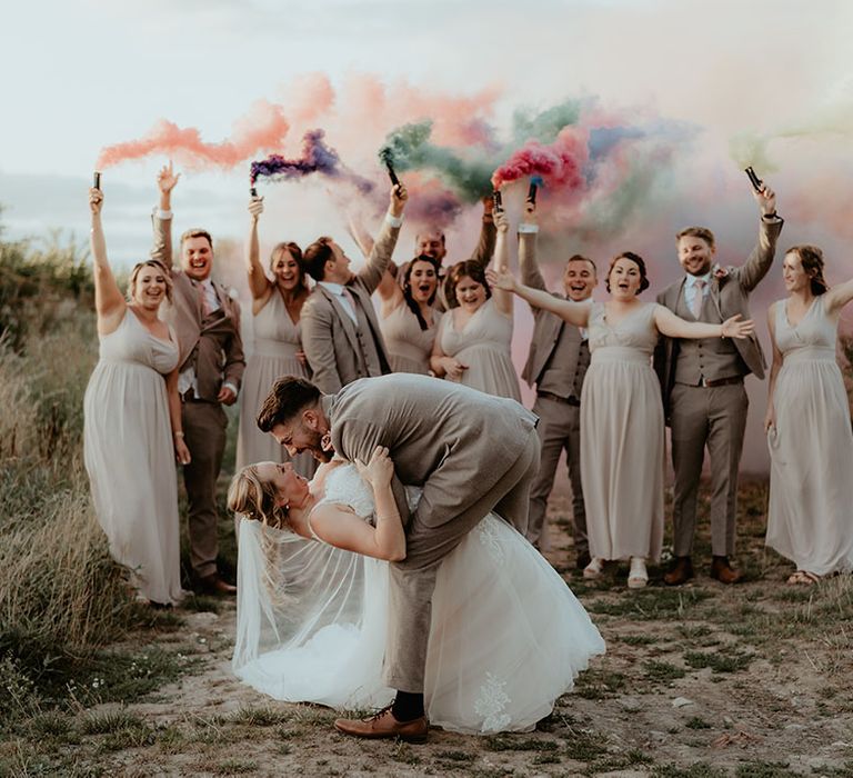 Bride & groom kiss surrounded by wedding party holding colourful smoke-bombs 