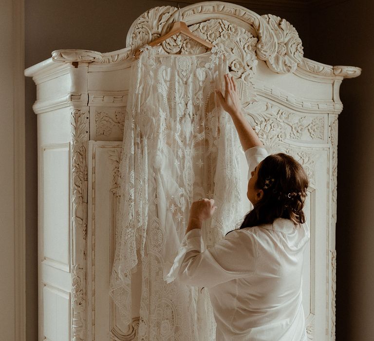 The bride in white satin pyjamas looks at her white lace boho wedding dress with her hair in a half up half down hairstyle with gold star accessories 