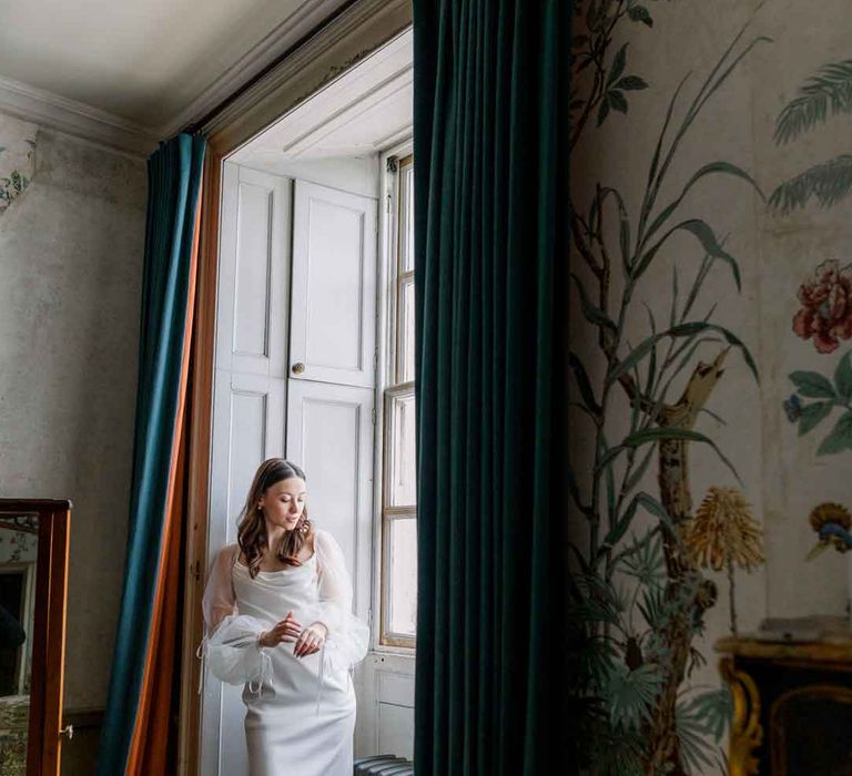 Bride in cowl neck satin wedding dress with sheer puff sleeves standing in the reception room of St Giles House 