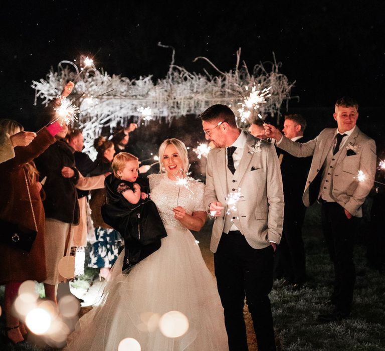 Bride and groom holding their little girl in a black dress and jacket waving their sparklers around at the end of the day