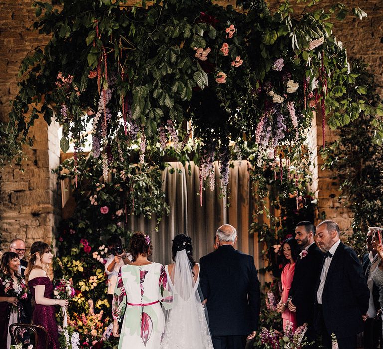 Extravagant purple and pink flower arrangements line the aisle and altar with candles in lanterns and the bride in a flower wedding dress being walked down the aisle by her parents