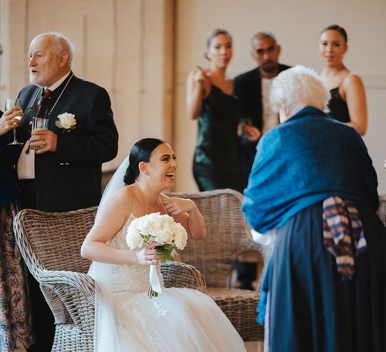 Bride lace sleeveless v neck Enzoani wedding dress with tulle finish and church length veil with 3d appliqué embroidered flowers sitting on a wicker chair at Grittleton House wedding venue holding white garden rose bouquet chatting with wedding guests 