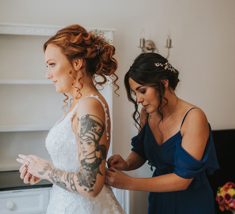 Bridesmaid in an off the shoulder dress helping the bride with tattoos into her lace princess wedding dress