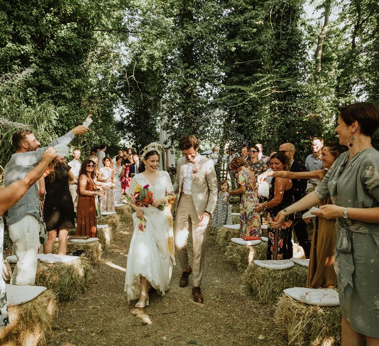 Bride in lace wedding dress and bridal crown leaves woodland wedding ceremony with groom in greige suit and colourful bow tie with guests cheering