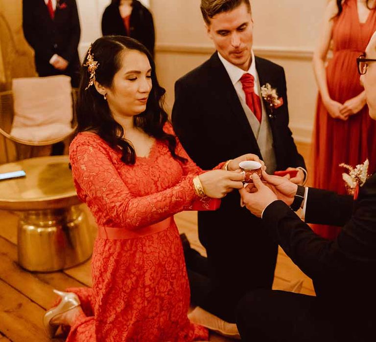 Groom in deep blue grooms blazer, grey waistcoat, red tie, red pocket square and mixed dried flower boutonniere and bride in red lace wedding dress with long sleeves, red and gold hair accessories and golden closed toe wedding heels holding teacup at Chinese Tea Ceremony 