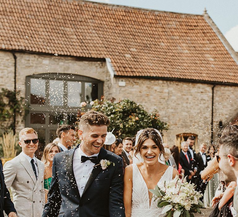 Groom in black tie walking with the bride in a fitted wedding dress to confetti 