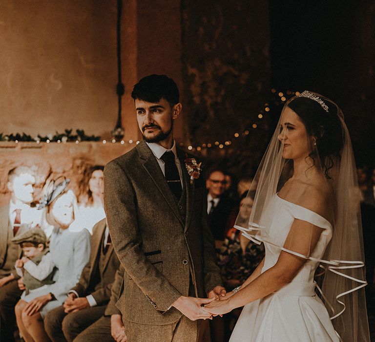 Groom in three piece grey suit with red tie and tie clip holds hands with the bride in an off the shoulder wedding dress and veil with sparkling tiara 