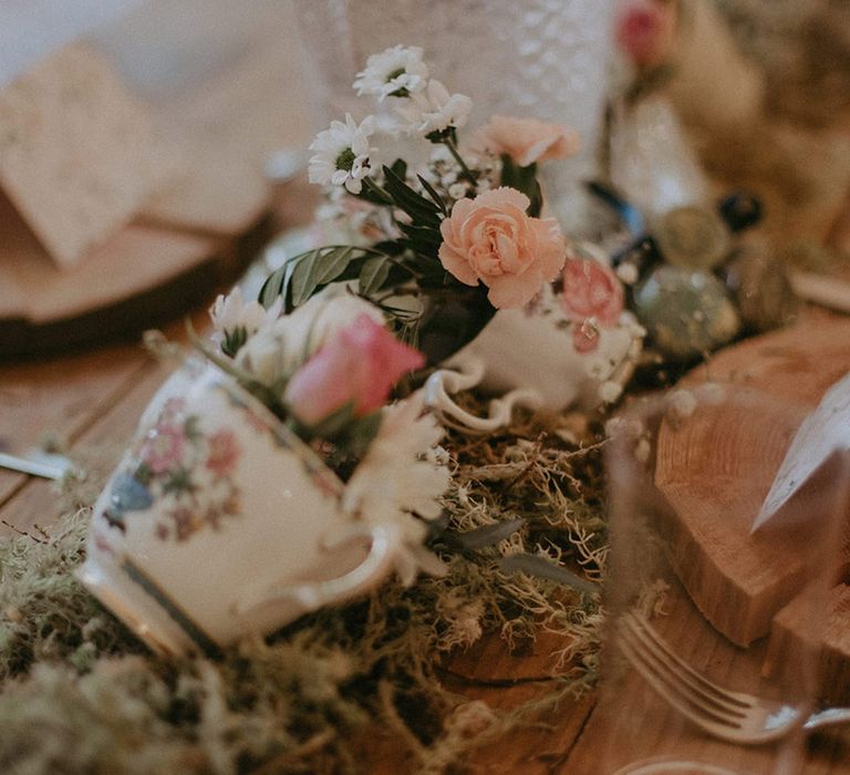 Teacups filled with pink flowers resting on bed of moss for the table decorations at boho woodland wedding 