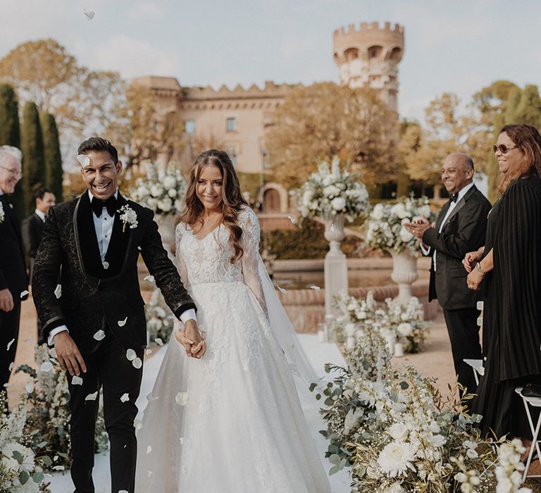 Bride in lace long sleeve Pronovias wedding dress and church-length veil with groom in classic black tuxedo with white flower boutonniere doing white petal confetti exit 