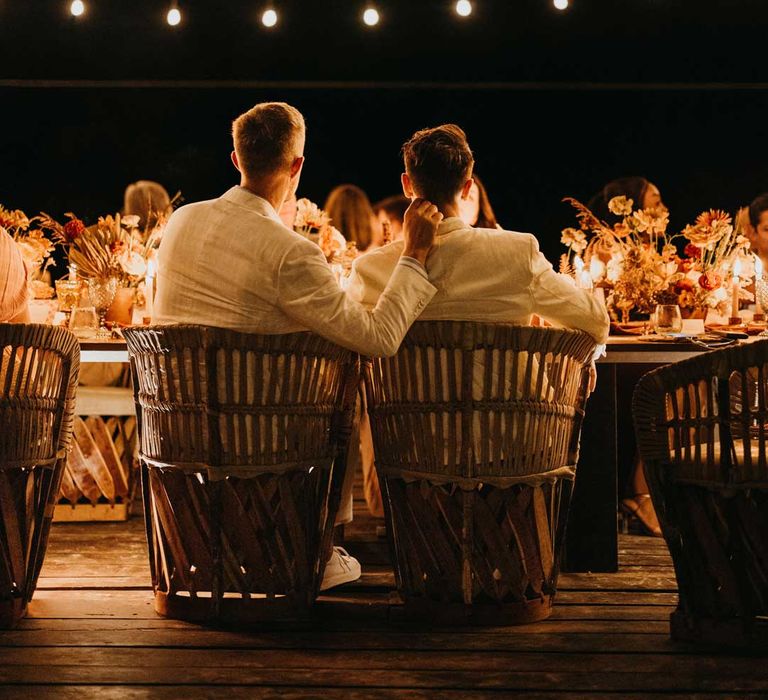 Grooms in complimenting white grooms wedding suits at wedding dinner under suspended fairy lights 