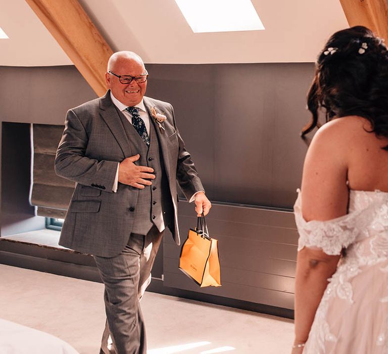 Father of the bride in grey three piece suit with floral tie walks in to see the bride in her gown for the first time carrying a gift bag 