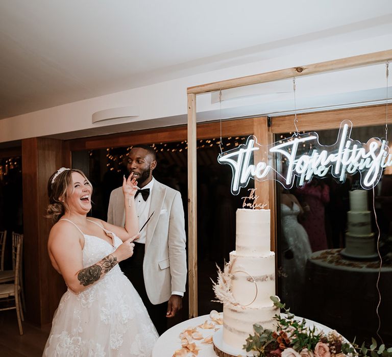 Three tier semi-naked rustic wedding cake with personalised cake topper sits in front of personalised neon sign ready to be cut by the bride and groom 