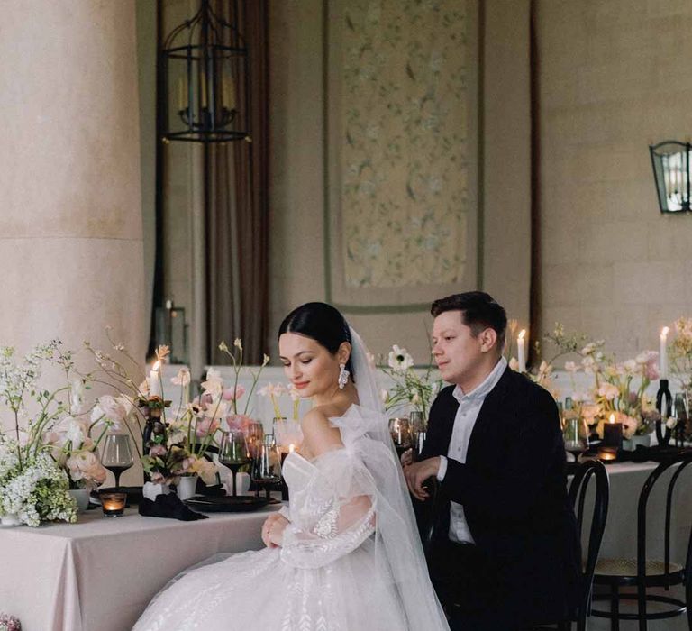 Bride in sweetheart neckline tulle mesh bridal gown with detachable puff tulle sleeves and delicate beading with cathedral length veil sitting with groom in classic black grooms suit at Notan inspired wedding tablescape at Settrington Orangery