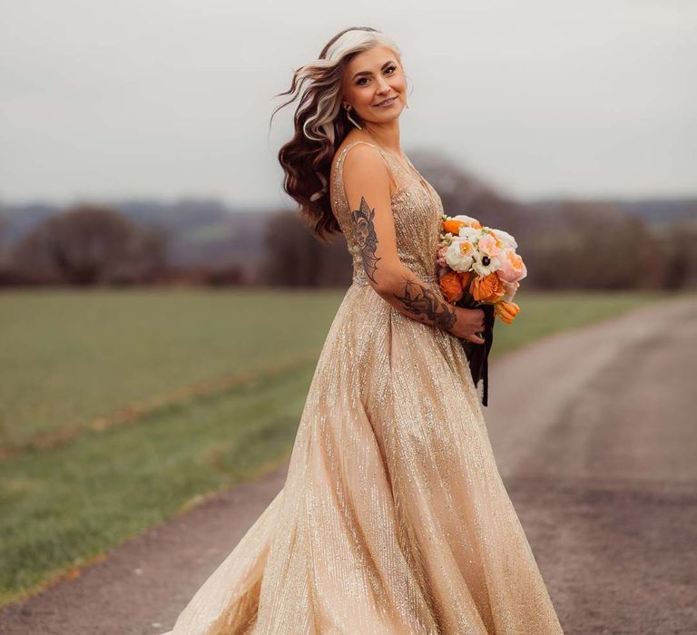 Bride spinning around wearing sleeveless deep v-neck sparkly gold wedding dress with orange garden rose, white poppy, light pink carnation and foliage bridal bouquet wrapped in sparkly gold ribbon at Kingscote Barn Tetbury 