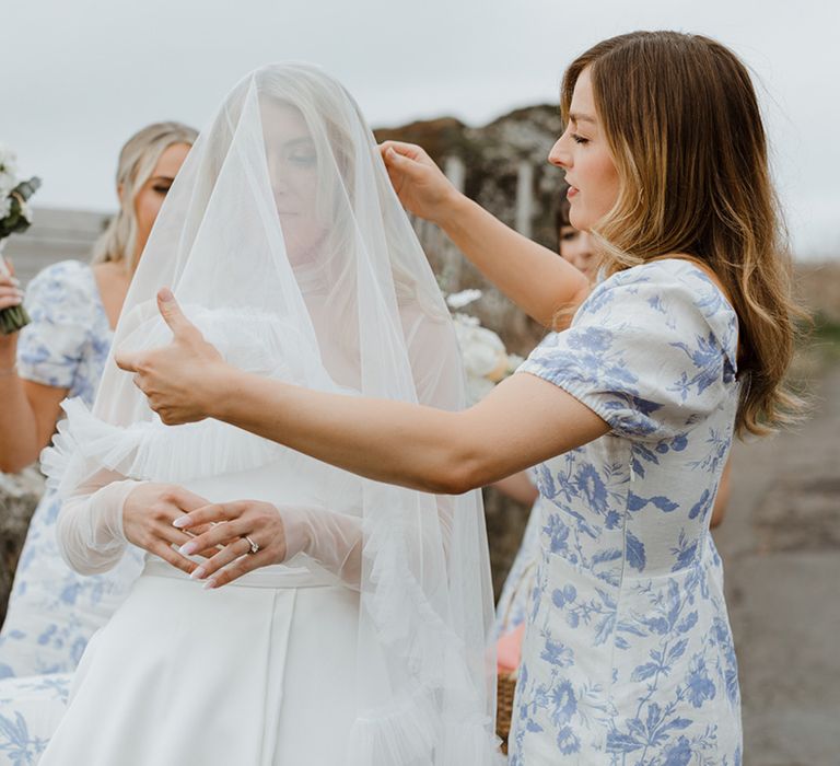 The bridesmaids in blue patterned bridesmaid dresses help the bride with her veil