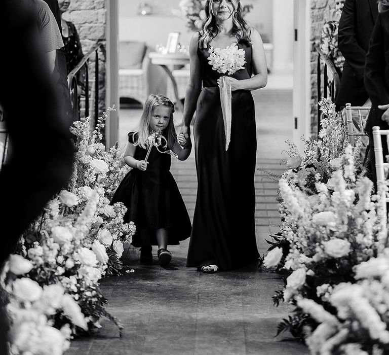 The bridesmaid and flower girl in navy dresses walk down the aisle holding hands 