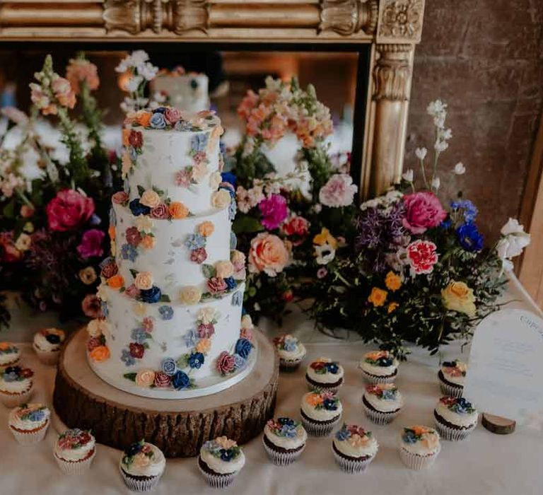 Three tiered iced wedding cake with spring flower icing decorations on wooden slate with wedding cupcakes with edible spring flowers