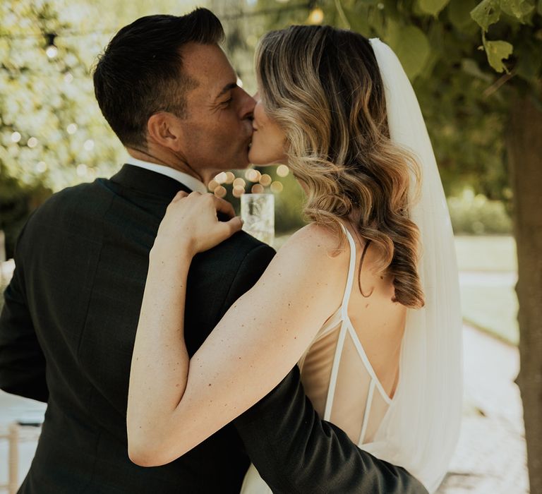 Bride wearing thin strap wedding dress with wedding veil and curled hair kissing the groom 