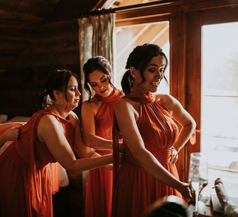 Bridesmaids wearing orange autumnal bridesmaid dresses helping each other all get ready for their day 