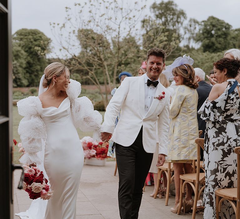 Bride in detachable puff pearl sleeves walking with groom in white tuxedo at their outdoor wedding ceremony 