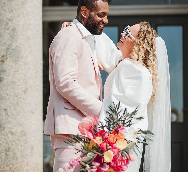 Groom in pastel pink wedding suit posing with the bride in a puff sleeve short wedding dress carrying hot pink wedding bouquet 